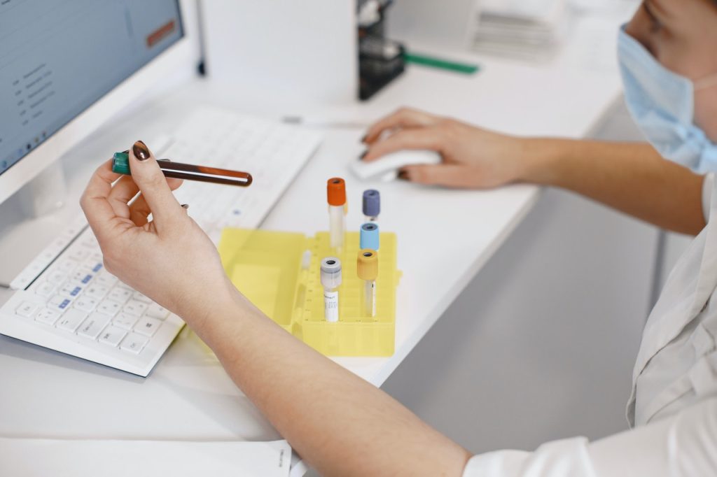 Masked laboratory technician holding a blood sample tube at a workstation