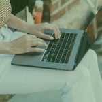 Person typing on a laptop outdoors beside a brick wall