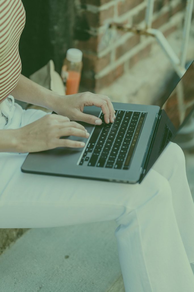 Person typing on a laptop outdoors beside a brick wall
